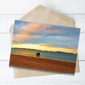 Greeting card featuring a romantic couple walking along Eastern Beach at sunset, with soft waves and golden sky in the background – photographed in New Zealand